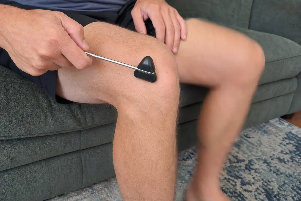 Närbild på en hand som testar knäreflexen med en reflexhammare i ett vardagsrum, så fungerar rörelsemönstren på vibrations...